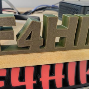 May include: A 3D-printed sign spelling out "E4HIK" in a blocky, sans-serif font. The letters are a muted olive green colour and sit on a light brown surface. A similar sign in red is visible below.