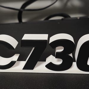 May include: A 3D-printed sign displaying the text "C7300" in black and white. The "C" has a white background with a black outline. A white and red circle is attached to the left side of the sign.