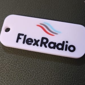 May include: A white rectangular keyring with rounded corners. The keyring has a hole on the left side. The text "FlexRadio" is printed in black, with a logo above it featuring red, grey, and blue curved lines.