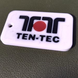 May include: A white rectangular keyring with rounded corners. The keyring features the black text "TEN-TEC" and a red circle. The top of the keyring has a small hole for attachment.