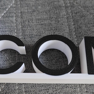 May include: A 3D-printed sign spelling "iCOM". The letters are white with black outlines, and the "i" has a red circle. The sign is mounted on a white base. The sign is likely for a business or personal use.