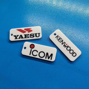 May include: Three white rectangular keychains with black text and logos. The keychains feature the brands "YAESU", "ICOM", and "KENWOOD". The "YAESU" keychain has a red logo. The "ICOM" keychain has a red dot. The "KENWOOD" keychain has a red dot.