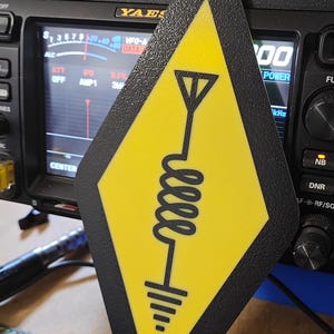 May include: A yellow and black diamond-shaped pendant with a stylized antenna symbol. The pendant is suspended in front of a radio receiver. The antenna symbol includes a coil, a vertical line, and ground lines.
