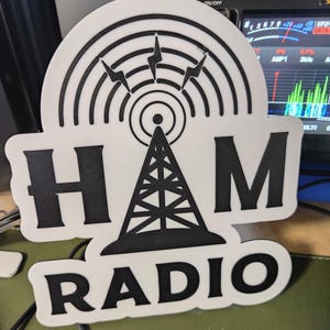 May include: A white and black sign featuring the text "HAM RADIO" and a graphic of a radio tower with signal lines and lightning bolts. The sign is likely for amateur radio enthusiasts.