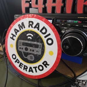 May include: A circular sign reading "HAM RADIO OPERATOR" in black lettering, with a red and white border. The sign displays a radio transceiver graphic and a signal icon. A black radio is visible in the background.