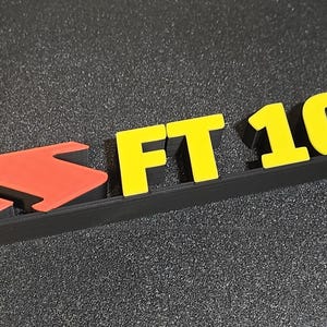 May include: A black and red arrow-shaped design with the text "FT 1000" in yellow. The letters and numbers are raised and have a 3D appearance. The design is set against a dark, textured background.