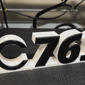 May include: A 3D-printed sign with the letters "LC7610" in black and white. A red and white circular object is attached to the left side. The sign is on a black surface.