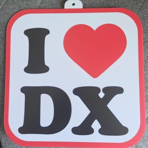 May include: Square sign with a white background, red border, and a red heart. The sign reads "I DX" in black block letters. The sign has a small hole at the top for hanging.