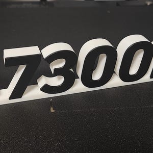 May include: A 3D-printed sign in white and black, with the text "IC7300MK2". The letters are black with a white outline, set on a white base. A red and white circular element is attached to the left.