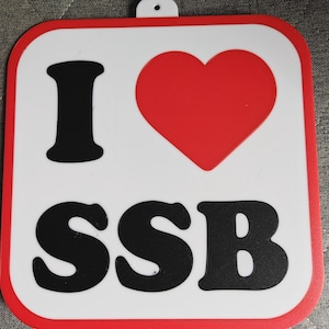May include: A white square trivet with a red border features a large red heart and the black letters "I" and "SSB". The trivet has a small hole at the top for hanging. The trivet is likely made of silicone or a similar heat-resistant material.