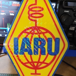 Peut inclure: Autocollant en forme de losange jaune et rouge avec les lettres IARU en bleu. L'autocollant présente un globe rouge, un éclair et un motif de bobine. L'arrière-plan comprend un équipement radio.