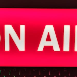 Op de afbeelding: Een rechthoekig, verlicht bord met de woorden "ON AIR" in vette, witte hoofdletters tegen een felrode achtergrond. Het bord wordt waarschijnlijk gebruikt in een opnamestudio of uitzendomgeving.