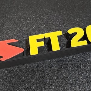 May include: A black and red emblem with the text "FT 2000" in yellow. The emblem features a red arrow-like shape on the left and raised letters and numbers. The background is a dark, textured surface.