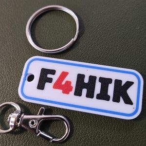 May include: A white rectangular keyring with the text "F4HIK" in black and red, outlined in blue. A silver key ring and a silver lobster clasp are also present, on a green surface.