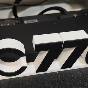 May include: A white and black 3D-printed sculpture with the text "IC7760". The sculpture includes a circular design with a red inner ring. The numbers and letters are in a bold, blocky font.