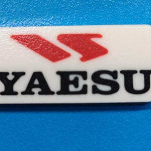 May include: A white rectangular keychain with rounded corners. The keychain features the word "YAESU" in black capital letters and a red arrow logo. A small hole is drilled in the upper left corner.