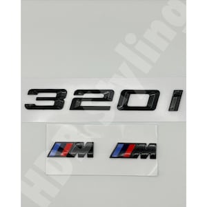 May include: A set of glossy black automotive emblems featuring the alphanumeric text 320i in a bold, modern font, accompanied by two smaller rectangular badges displaying the letter M with blue, purple, and red diagonal stripes on a white backing.