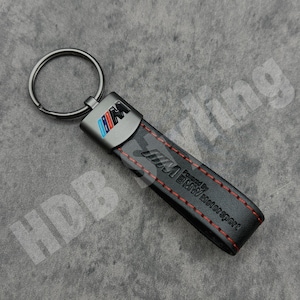 May include: A black leather keyring featuring a metal attachment with the BMW M logo in blue, purple, and red. The leather strap is embossed with the BMW Motorsport text and logo, accented by red stitching, and includes a circular metal ring.
