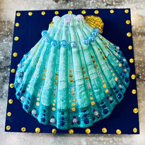 Könnte beinhalten: Ein dekoratives Muschelkunstwerk. Die Muschel ist in Türkis- und Blautönen bemalt, mit goldenen Akzenten und einer Reihe blauer Perlen verziert. Sie ist auf einem quadratischen, marineblauen Hintergrund mit goldenen Punkten an den Rändern montiert.