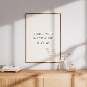 May include: A framed print with the text "You're allowed to outgrow what no longer fits" in a brown font. The print is displayed on a wooden cabinet with a vase and a small glass bottle. A small wooden tray with books is on the cabinet.