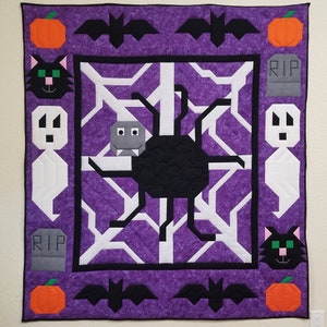May include: A purple quilted wall hanging with a black spider web design. The web has a black spider in the center. The border of the quilt is decorated with black bats, orange pumpkins, white ghosts, black cats, and gray tombstones with the word "RIP" on them.