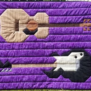 May include: A purple quilted fabric wall hanging with two appliqued guitars. The top guitar is beige with brown accents and the bottom guitar is black and white.
