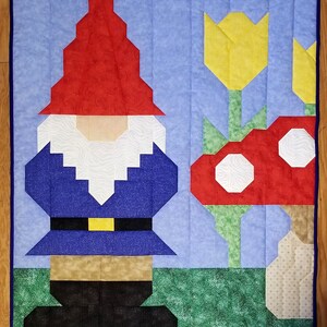Könnte beinhalten: Ein blauer und roter Patchwork-Zwerg mit einem gelben Gürtel steht vor einem roten und weißen Pilz und einer gelben Tulpe auf blauem Hintergrund.