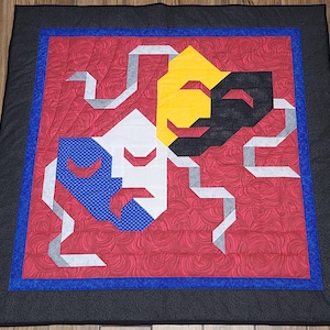 Puede incluir: Un edredón hecho a mano con cuatro máscaras de teatro estilizadas en blanco, amarillo, negro y azul, sobre un fondo rojo. El edredón tiene un borde azul, un borde gris y un borde negro.