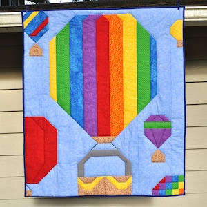 Könnte beinhalten: Eine bunte Patchwork-Decke mit einem großen Heißluftballon mit Regenbogenstreifen. Der Ballon ist von kleineren Heißluftballons in verschiedenen Farben und Mustern umgeben. Die Decke ist aus Baumwollstoff und perfekt für ein Kinderzimmer oder ein Babyzimmer.