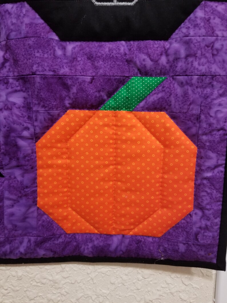 May include: A purple and orange patchwork quilt block featuring a pumpkin design. The pumpkin is orange with white polka dots and has a green stem with white polka dots.
