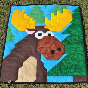 Puede incluir: Un alce marrón con cuernos amarillos y ojos marrones está rodeado de árboles verdes y azules. El alce está hecho de tela y es parte de una colcha.