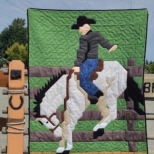 Puede incluir: Un tapiz de pared acolchado que representa a un vaquero montando un potro salvaje. El edredón presenta un fondo verde texturizado, una valla marrón y un jinete con vaqueros azules y sombrero negro. El caballo es blanco y marrón.