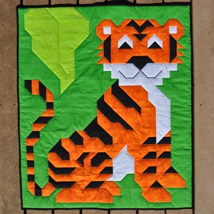May include: A green fabric square with a white and orange tiger applique. The tiger is sitting with its tail curled behind it. The tiger has black stripes and a white face.