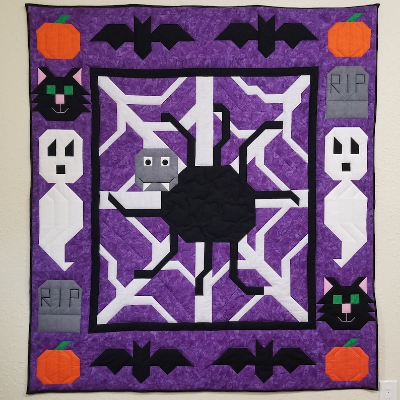 May include: A purple quilted wall hanging with a black spider web design. The web has a black spider in the centre. The border of the quilt is decorated with black bats, orange pumpkins, white ghosts, black cats, and grey tombstones with the word "RIP" on them.