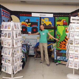 May include: A woman stands in front of a booth displaying counted cross stitch patterns. The booth is decorated with colorful quilts featuring various themes, including a monster truck, construction vehicles, and a tiger. The sign above the patterns reads "Counted Quilts".
