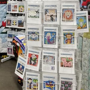 May include: A white metal display stand with multiple rows of colorful printed fabric squares. Each square features a different design, including animals, people, and objects. The designs are all in a pixelated style, reminiscent of cross-stitch or embroidery.