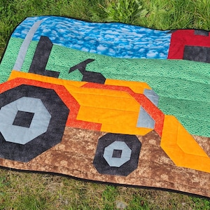 Op de afbeelding: Een kleurrijke quilt met een tractorontwerp. De quilt toont een oranje tractor met zwarte en grijze wielen, tegen een achtergrond van een groen veld, blauwe lucht en een rode schuur. De quilt ligt op groen gras.