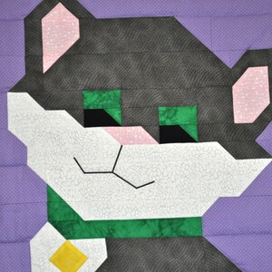 Op de afbeelding: Een patchwork quiltblok met een gestileerde kat gemaakt van zwarte, witte, groene en roze stof. De kat heeft groene ogen en een wit gezicht.