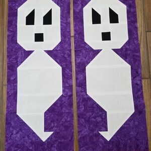 May include: Two white fabric ghost shapes with black facial features on a purple background. The ghosts are cut out and ready to be sewn together.