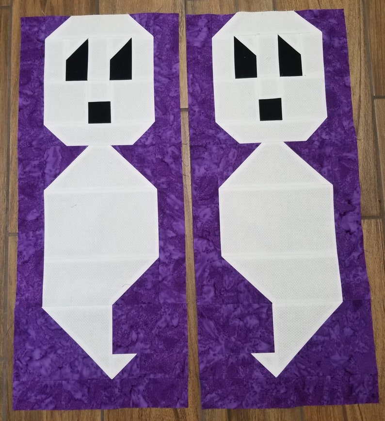May include: Two white fabric ghost shapes with black facial features on a purple background. The ghosts are cut out and ready to be sewn together.