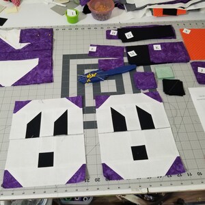 May include: A close-up of a quilting project in progress. Two white squares with black geometric shapes are surrounded by purple fabric squares. The squares are on a cutting mat with grid lines. There are various other fabric pieces and tools scattered around the mat.