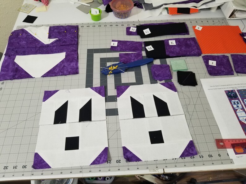 May include: A close-up of a quilting project in progress. Two white squares with black geometric shapes are surrounded by purple fabric squares. The squares are on a cutting mat with grid lines. There are various other fabric pieces and tools scattered around the mat.