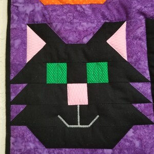 May include: A black cat face with green eyes and a pink nose, stitched onto a purple fabric background.