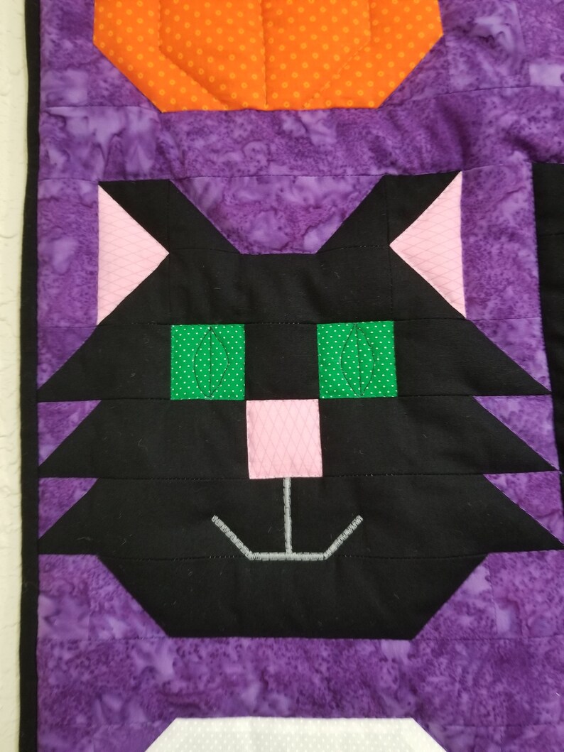 May include: A black cat face with green eyes and a pink nose, stitched onto a purple fabric background.