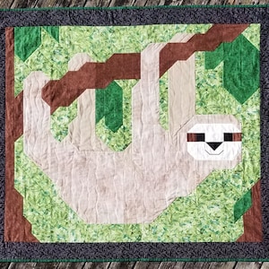 May include: A brown, green, and white patchwork quilt featuring a sloth hanging from a branch. The sloth is made of beige fabric and has a white face with black eyes.
