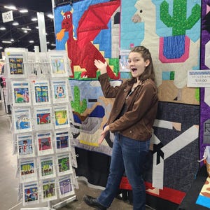 May include: A woman stands in front of a display of colorful quilt patterns. The patterns feature a dragon, a llama, and a cactus. The text "All Quilt Patterns $10.00" is visible on the display.