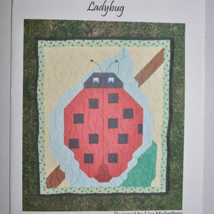 May include: A counted cross stitch pattern for a quilt featuring a red ladybug with black spots on a green leaf. The pattern is titled "Ladybug" and is designed by Lisa Muilenburg and quilted by Laneth Dick.