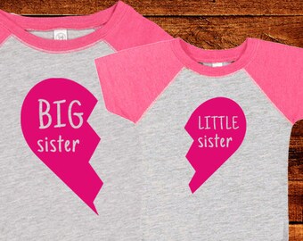 May include: Two shirts with a pink and gray color scheme. The larger shirt says "BIG sister" with a pink heart that is broken in half. The smaller shirt says "LITTLE sister" with the other half of the pink heart.