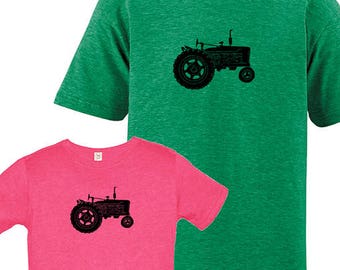 May include: A set of two t-shirts featuring a black silhouette of a vintage tractor. The adult t-shirt is green and the youth t-shirt is pink.