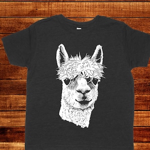May include: Dark gray t-shirt featuring a white illustration of an alpaca's face. The alpaca has a stylish haircut and is looking forward. The shirt is made of a soft material.